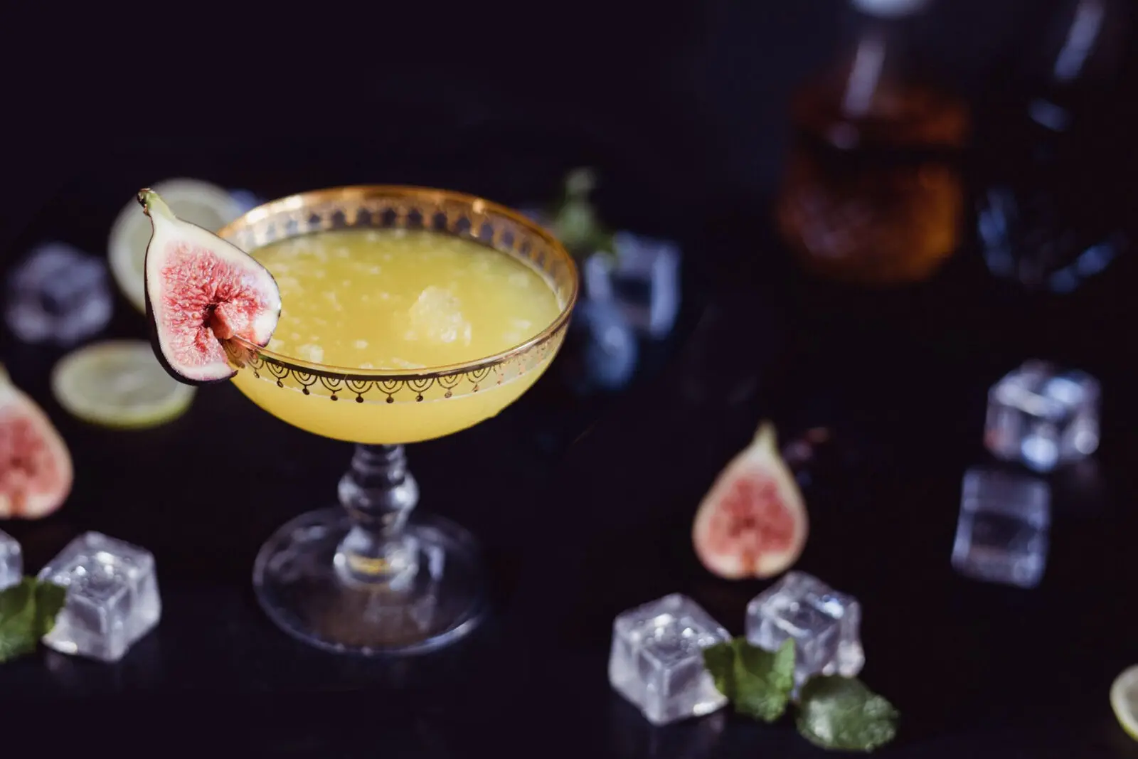 Cocktail à la figue et citron vert Un verre de cocktail élégant, décoré d'une tranche de figue, entouré de glaçons, de quartiers de citron vert et de morceaux de figue, sur un fond sombre.