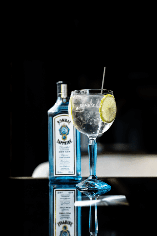 Verre de gin tonic Bombay Sapphire avec une tranche de citron, sur une table noire réfléchissante, accompagné de sa bouteille en arrière-plan.