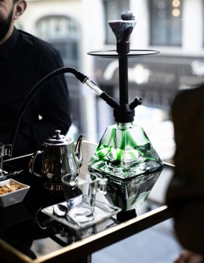 Une chicha moderne avec une base en verre vert, posée sur une table élégante avec une théière argentée et des accessoires, avec des personnes en arrière-plan.