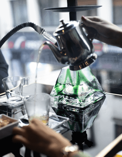 Une chicha en verre vert posée sur une table en verre noir réfléchissante, avec une main qui verse de l'eau à l'aide d'une théière en métal.