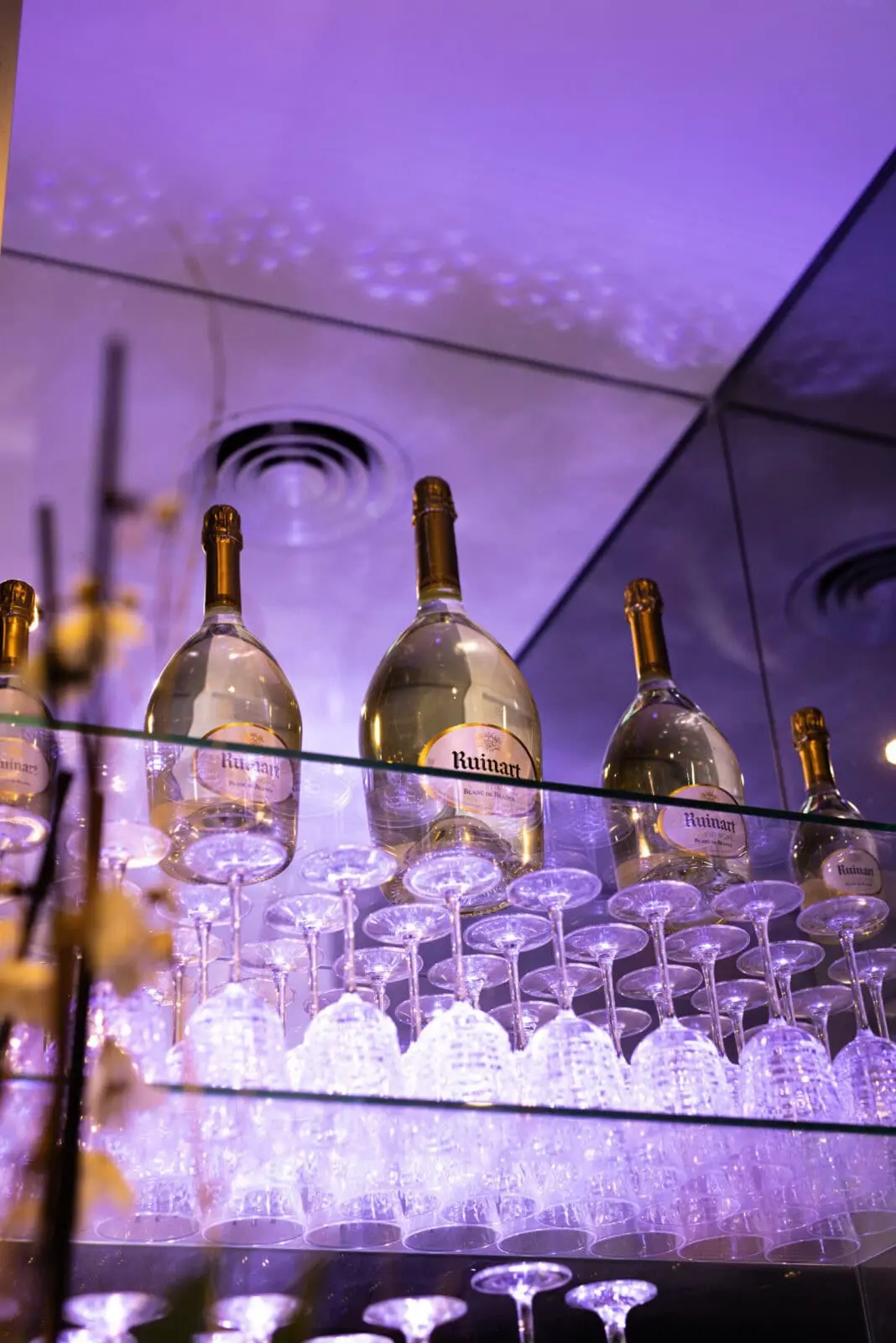 Présentoir de bouteilles de champagne et verres en cristal sous un éclairage violet Présentoir avec des bouteilles de champagne dorées et des verres en cristal, éclairé par une lumière violette dans un décor chic et moderne.